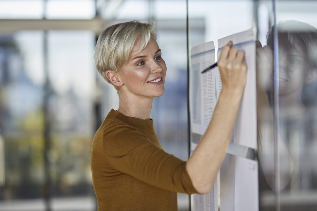 smiling businesswoman writing on papers at glass pane e1656541647888